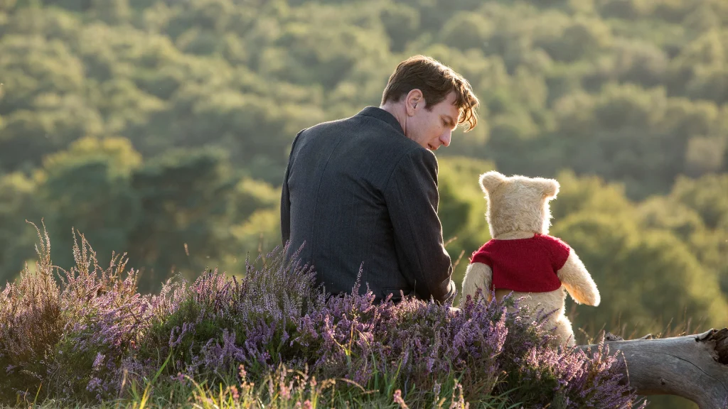 A man in a suit sits on grass next to a stuffed bear wearing a red shirt, overlooking a landscape with dense green trees.