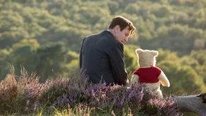 A man in a suit sits on grass next to a stuffed bear wearing a red shirt, overlooking a landscape with dense green trees.