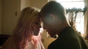 Two people with their foreheads touching in a softly lit room, sharing an intimate moment. The person on the left has light pink hair, and the person on the right has short hair.