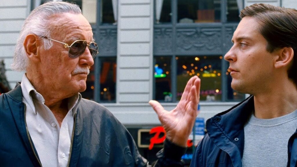 Two men standing outside, one wearing sunglasses and a jacket, the other gesturing with one hand. Building storefronts are visible in the background.