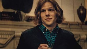 Person with shoulder-length hair holds a drink and is wearing a blue leopard-print shirt under a dark jacket, standing indoors with a neutral expression.