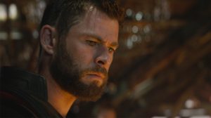 A bearded man with short hair is looking down with a serious expression in a dimly lit setting.