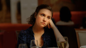A woman with a contemplative expression rests her head on her hand while seated at a dining table with a glass of white wine in front of her.