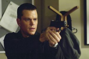 A man in a dark jacket pointing a handgun, standing indoors with a coat rack and bulletin board in the background.