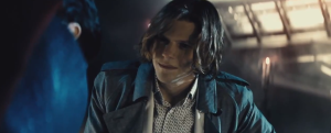 A person with long hair and a trench coat looks intently at someone in front of them, under subdued lighting.