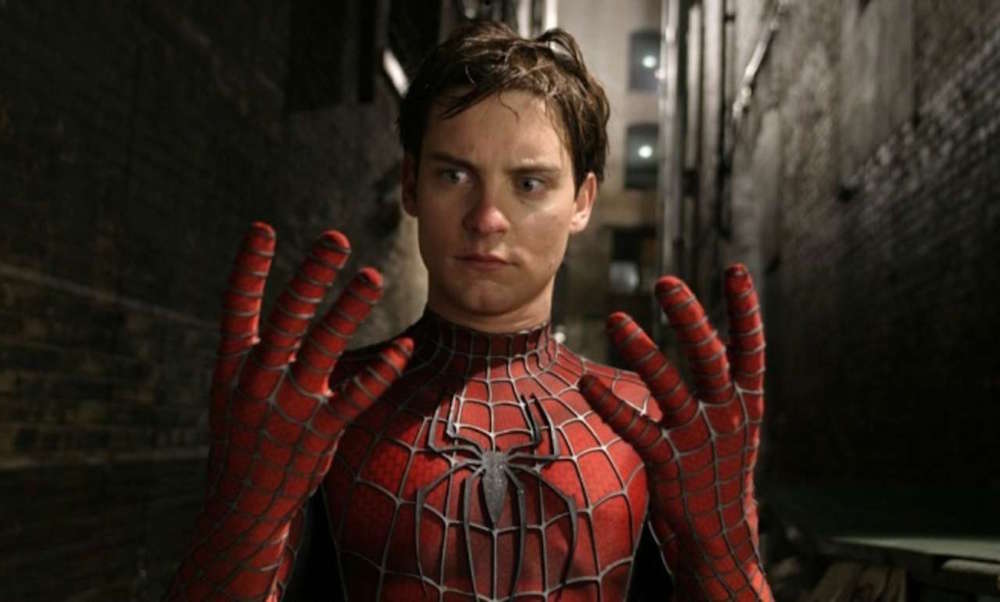 A man in a Spider-Man costume looks surprised with his hands raised in front of him, standing in a dimly lit alley.