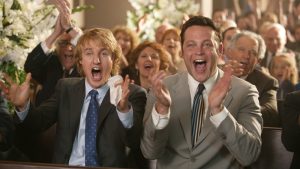 Director David Dobkin Talks Potential Sequel To ‘WEDDING CRASHERS’ Two men smiling and clapping enthusiastically in a crowd at a formal event, surrounded by other attendees.