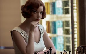 A woman with short, wavy red hair sits at a chessboard, contemplating her next move. She is wearing a white sleeveless dress and looking directly at the camera. A bookshelf is visible in the background.