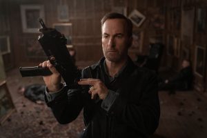 A man in a dark room holds and inspects a firearm. The background has framed pictures, scattered debris, and a person lying on the floor.