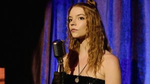 A woman with long, wavy hair stands on stage with a retro microphone in front of a blue curtain.