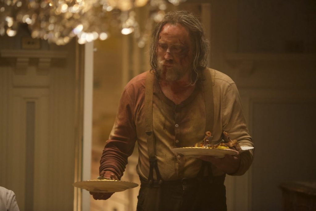 A person with disheveled hair and a stained shirt holds two plates of food in a dimly lit room.