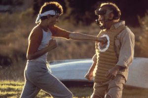 The Karate Kid Trilogy Is Heading To 4K UHD Blu-Ray This December Just In Time For New Season Of ‘Cobra Kai’ A young man wearing a headband punches a padded target held by an older man in protective gear outdoors.