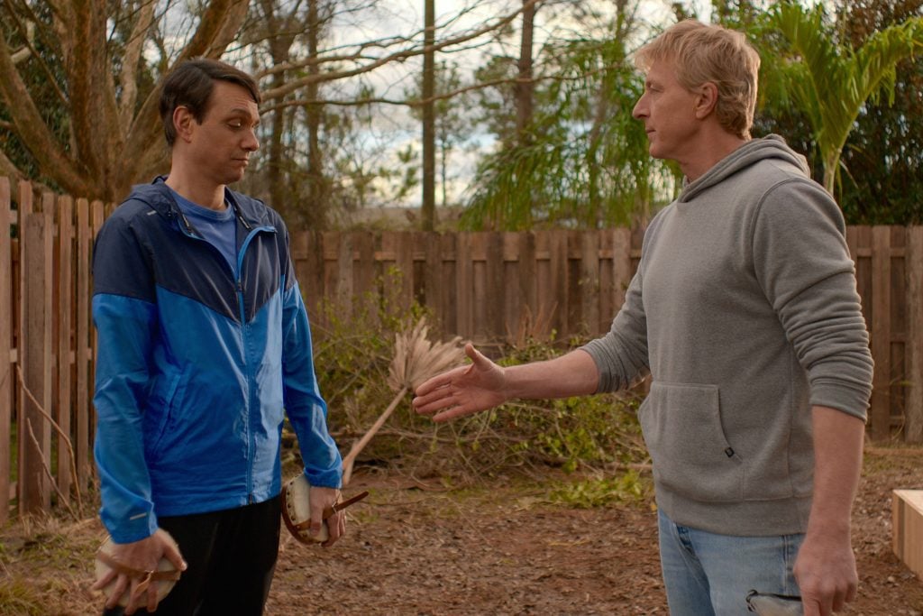 Two men are in a backyard. The man on the left, wearing a blue jacket, holds gardening tools and looks at the man on the right, who is extending a handshake. There is a wooden fence and trees in the background.