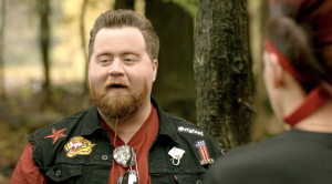 Man with a beard and long goatee wearing a patched vest talks to someone outdoors, with trees in the background.