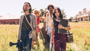 Four people carrying equipment and bags walk through a grassy field toward a farmhouse on a sunny day.
