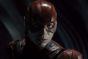 A person in a detailed red superhero suit and mask, with a serious expression, stands against a dark blurred background.