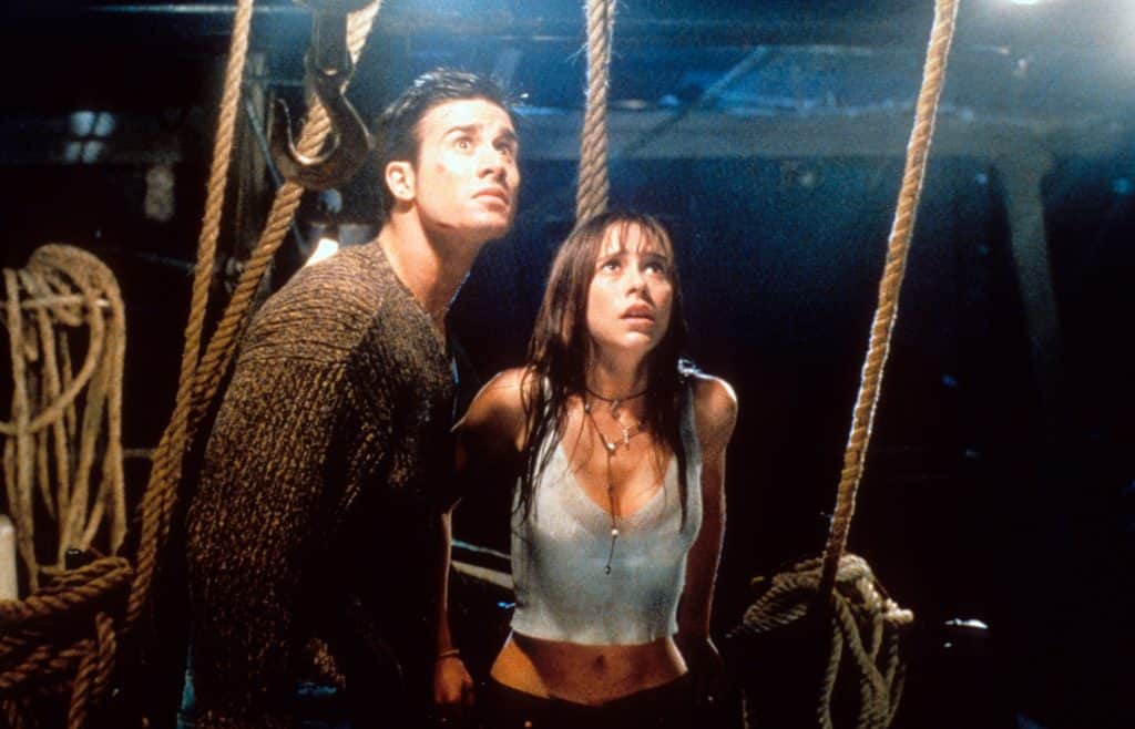 Two people on a dimly lit boat deck look upwards with tense expressions. The man wears a brown sweater, and the woman wears a light-colored tank top. Ropes and rigging are visible around them.