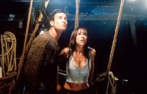 Two people on a dimly lit boat deck look upwards with tense expressions. The man wears a brown sweater, and the woman wears a light-colored tank top. Ropes and rigging are visible around them.