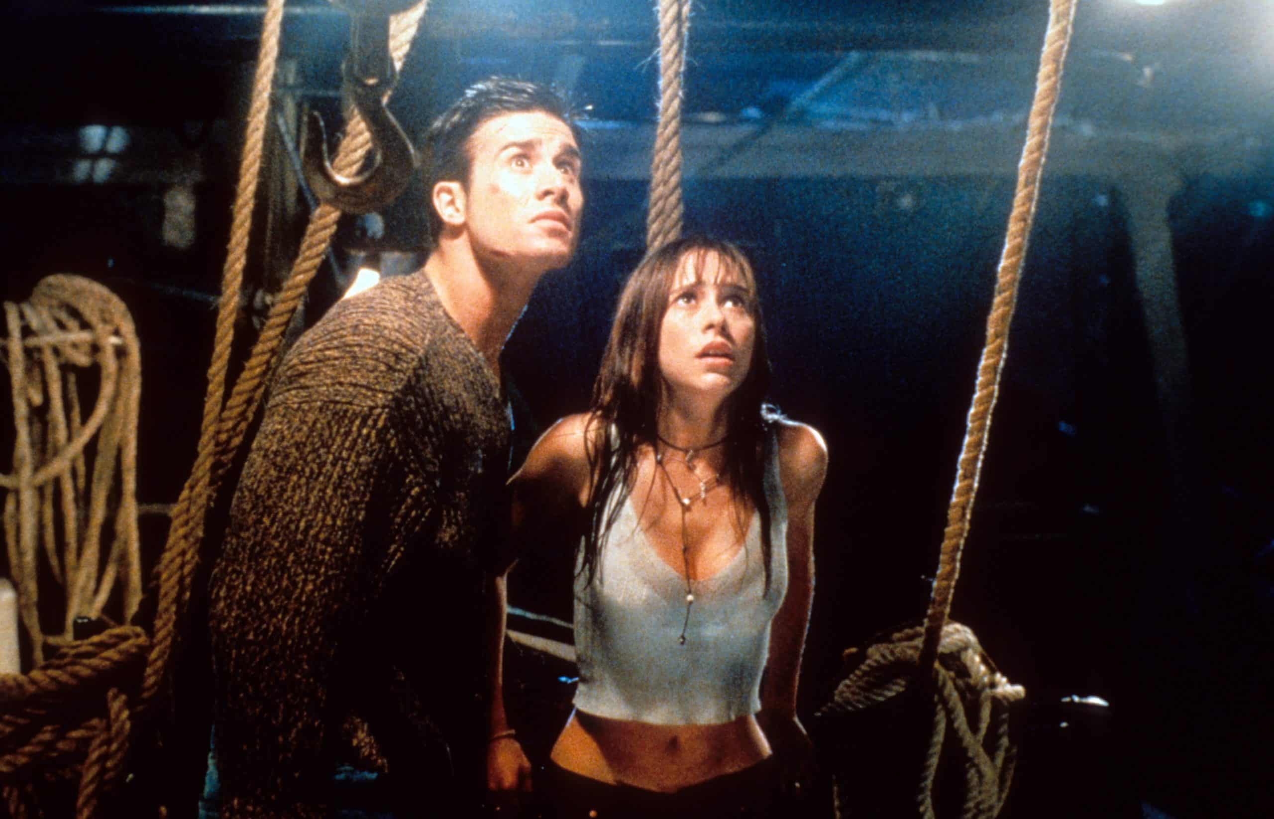 Two people on a dimly lit boat deck look upwards with tense expressions. The man wears a brown sweater, and the woman wears a light-colored tank top. Ropes and rigging are visible around them.