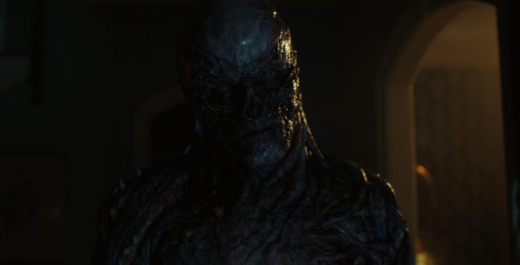 A dark, grotesque creature with a skeletal appearance stands in a dimly lit room, partially illuminated.