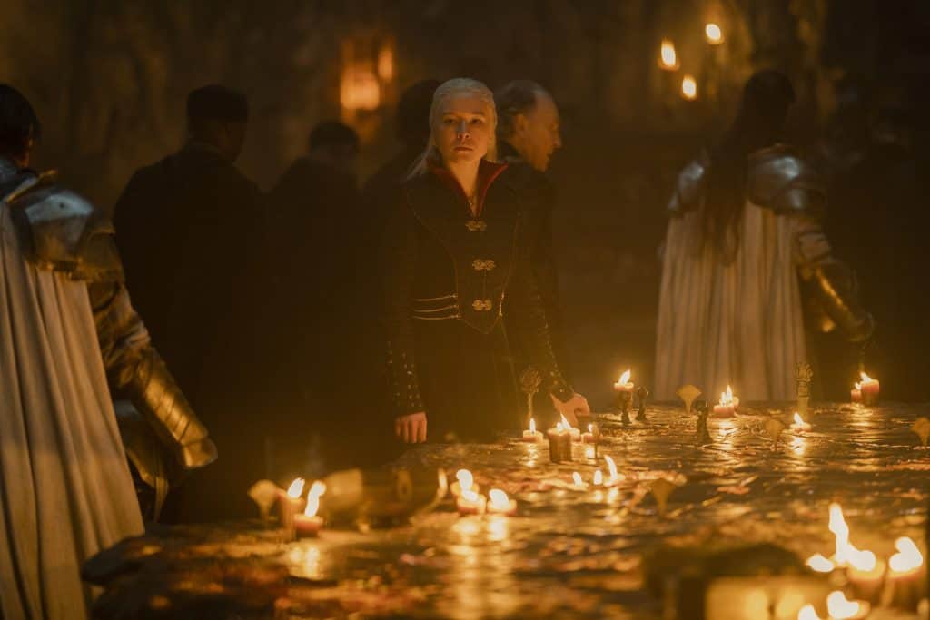 A person in dark medieval-style clothing stands by a candlelit table with maps and scrolls, surrounded by others in similar attire, in a dimly lit room.
