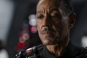 A man with a serious expression, wearing a black uniform with a badge, is seen in a close-up shot. The background is blurred with red and white lights.