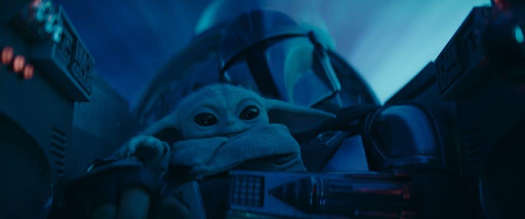 A small, green, big-eyed creature sits beside a helmeted figure in armor in a dark, blue-tinged environment.