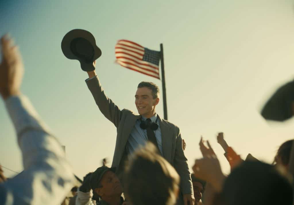Man in suit raising a hat with a crowd and american flag in the background.
