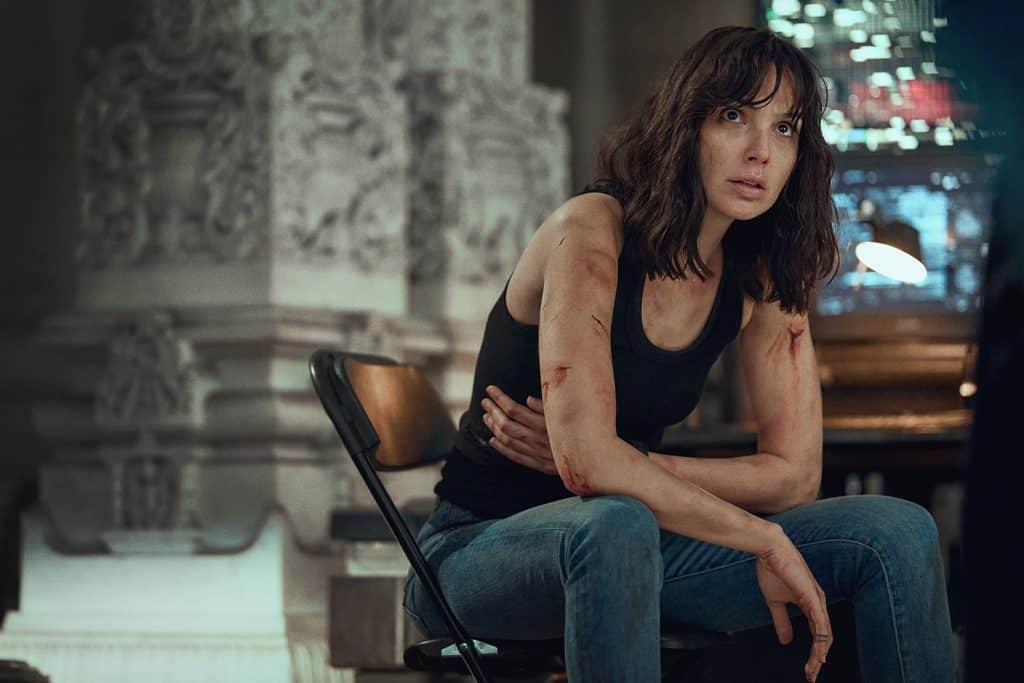A woman with disheveled hair and bruises, wearing a black tank top and jeans, sits on a folding chair in a dimly lit, ornate room, looking ahead with a tense expression.