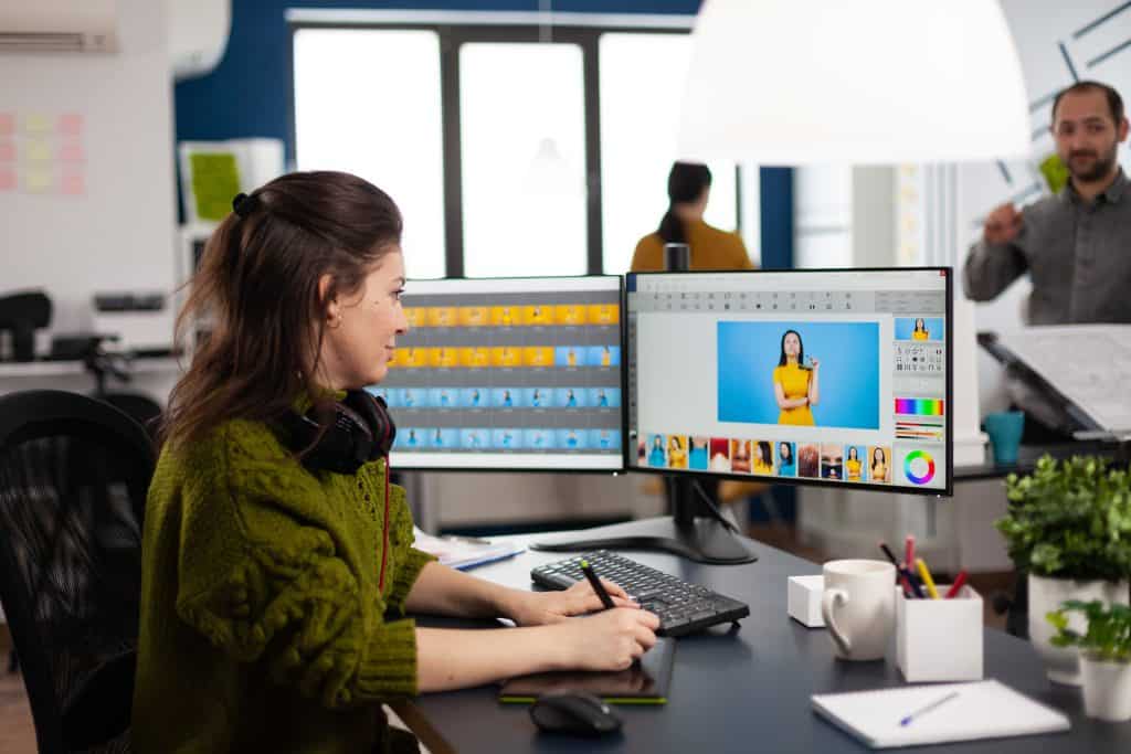 A woman working on a graphic design project on a computer in a busy office, with colleagues visible in the background.