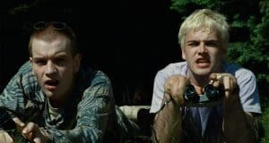 Two young men lie on the grass; one holds binoculars and a cigarette, both looking intently ahead, with trees in the background.