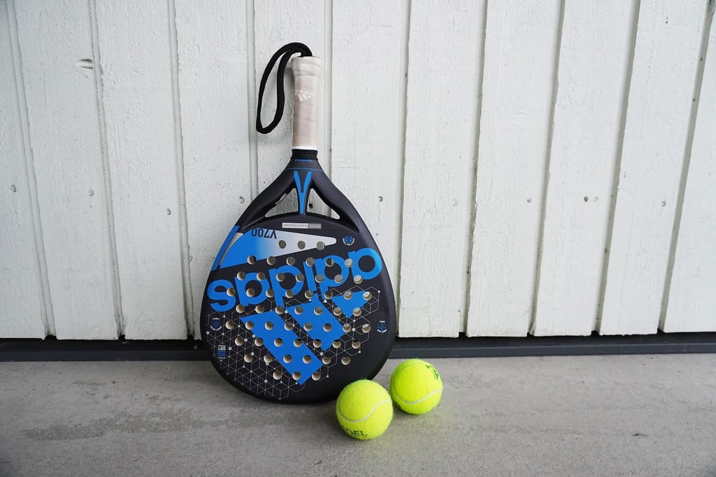 A tennis racket and two tennis balls next to a wall.