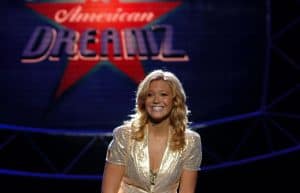 A woman in a gold dress sitting on a stage in front of an american dreamz sign.