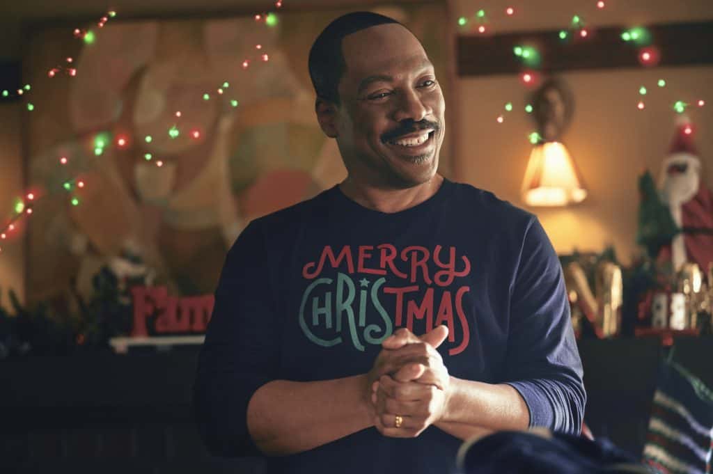 A man wearing a merry christmas shirt in front of a christmas tree.
