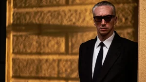 A man in a suit and sunglasses leaning against a wall.