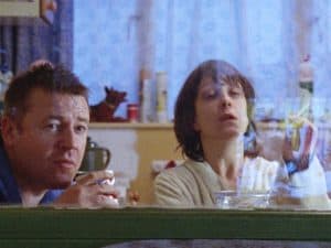 ‘Nil By Mouth’ Blu-Ray Review – Gary Oldman’s Directorial Debut Is A Soul-Crushing Endeavor A man and woman sitting in a kitchen looking through a window.