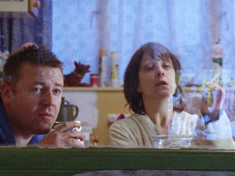 ‘Nil By Mouth’ Blu-Ray Review – Gary Oldman’s Directorial Debut Is A Soul-Crushing Endeavor A man and woman sitting in a kitchen looking through a window.