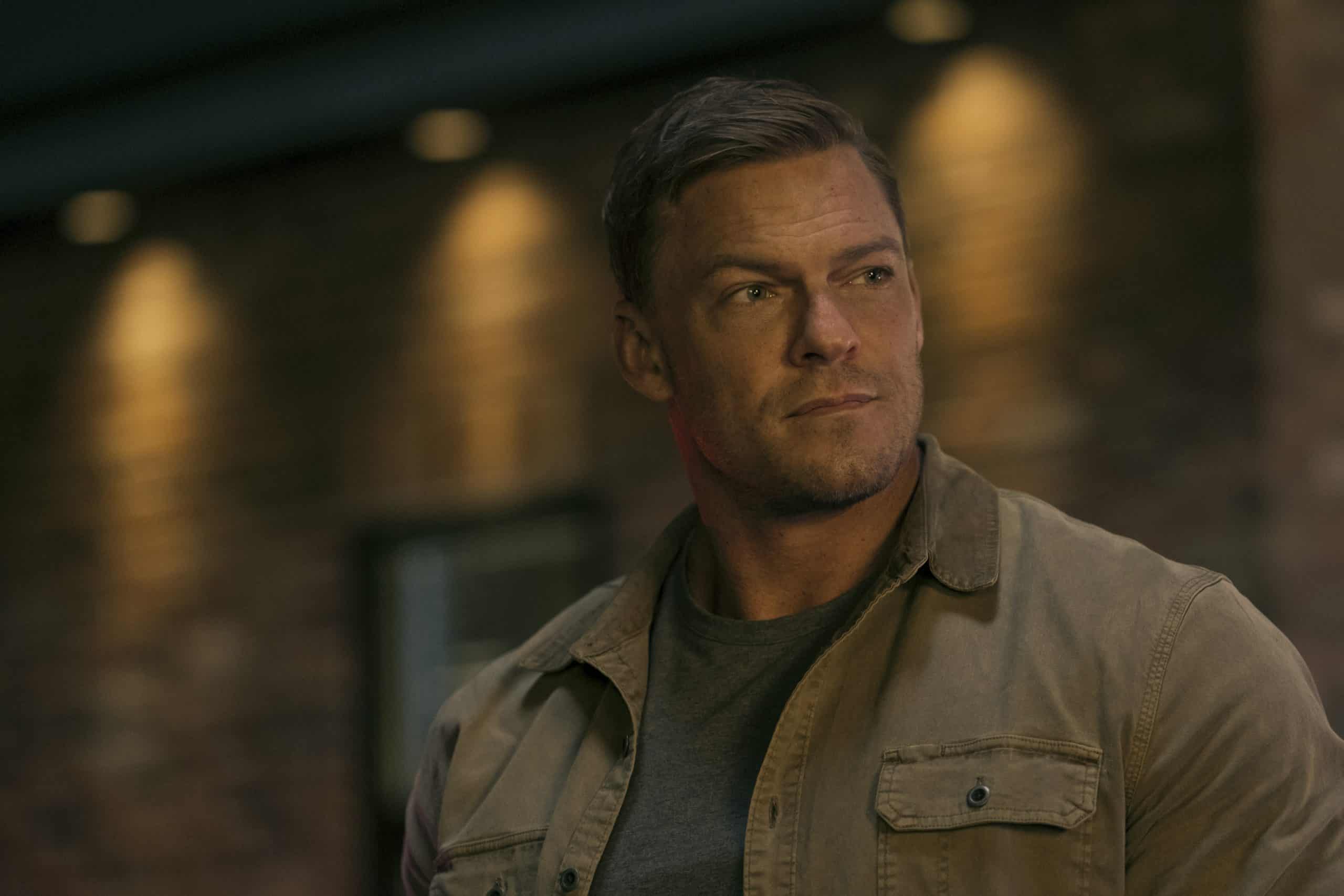 A man in a tan jacket standing in front of a brick wall.