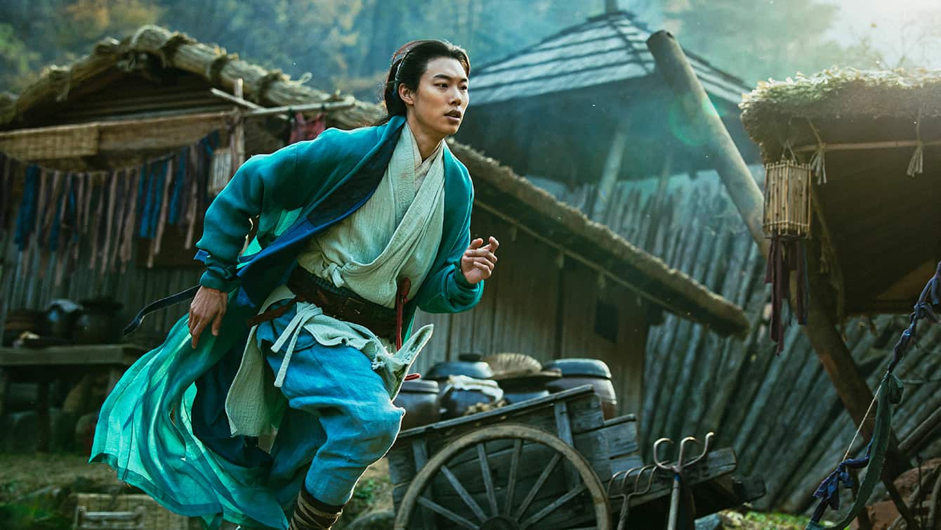 A man in a blue dress is running in front of a hut.