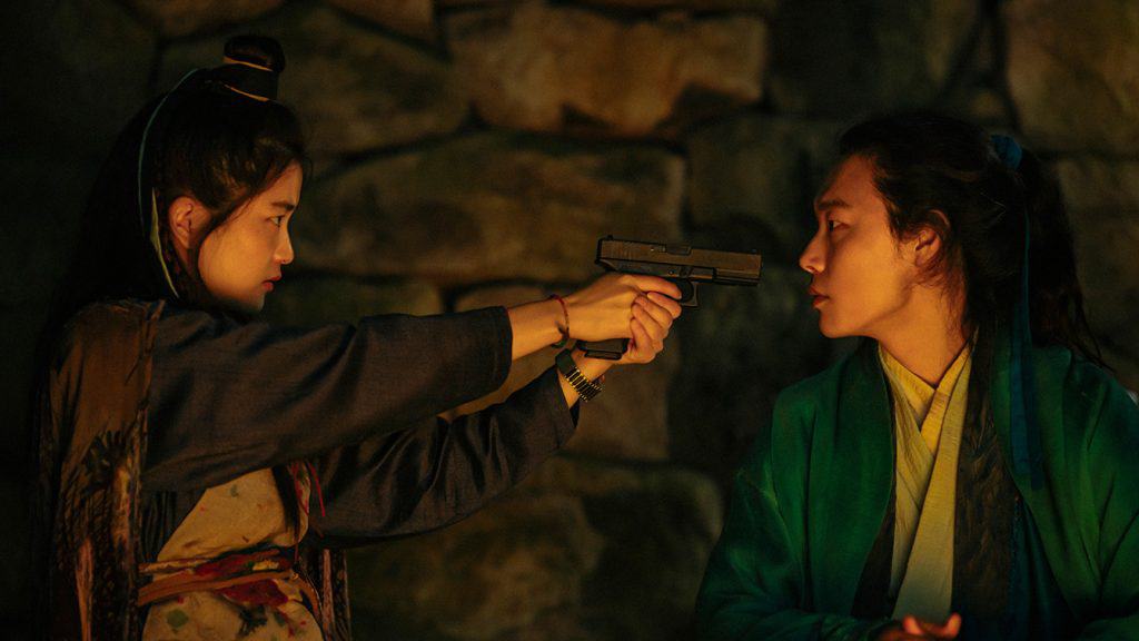 Two women are holding a gun in front of a stone wall.