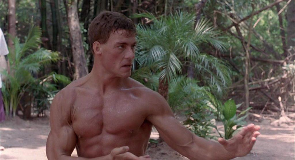 A shirtless man is doing martial arts in a wooded area.