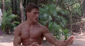 A shirtless man is doing martial arts in a wooded area.
