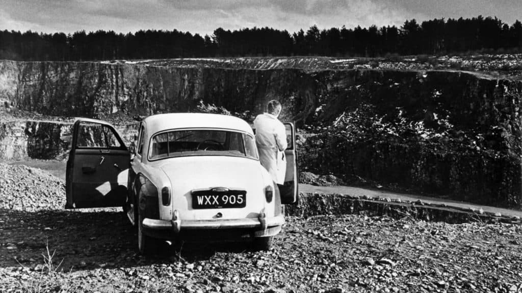 A black and white photo of a man standing next to a car.