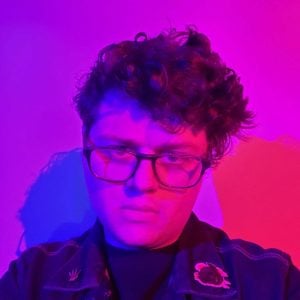 A man in glasses is standing in front of a purple and pink wall.