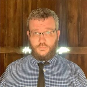 A man with glasses and a tie in front of a barn.