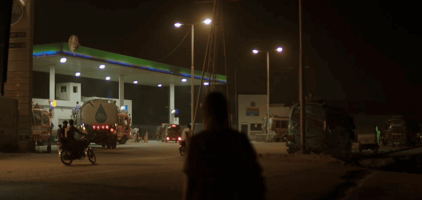 A nighttime scene of a gas station with vehicles and a person in the foreground.