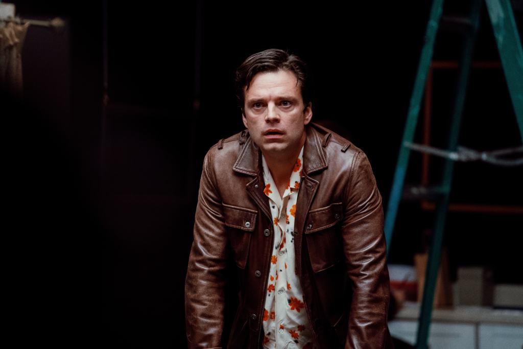 A man with a concerned expression wearing a brown leather jacket and a floral shirt, with a ladder in the background.