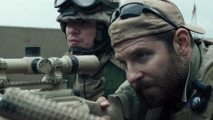 Two military personnel in combat gear with communication headsets operating a mounted weapon system.