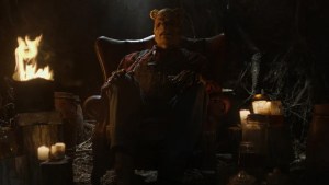 A costumed character sitting on a chair in a dimly lit setting with candles and a fire in the background.