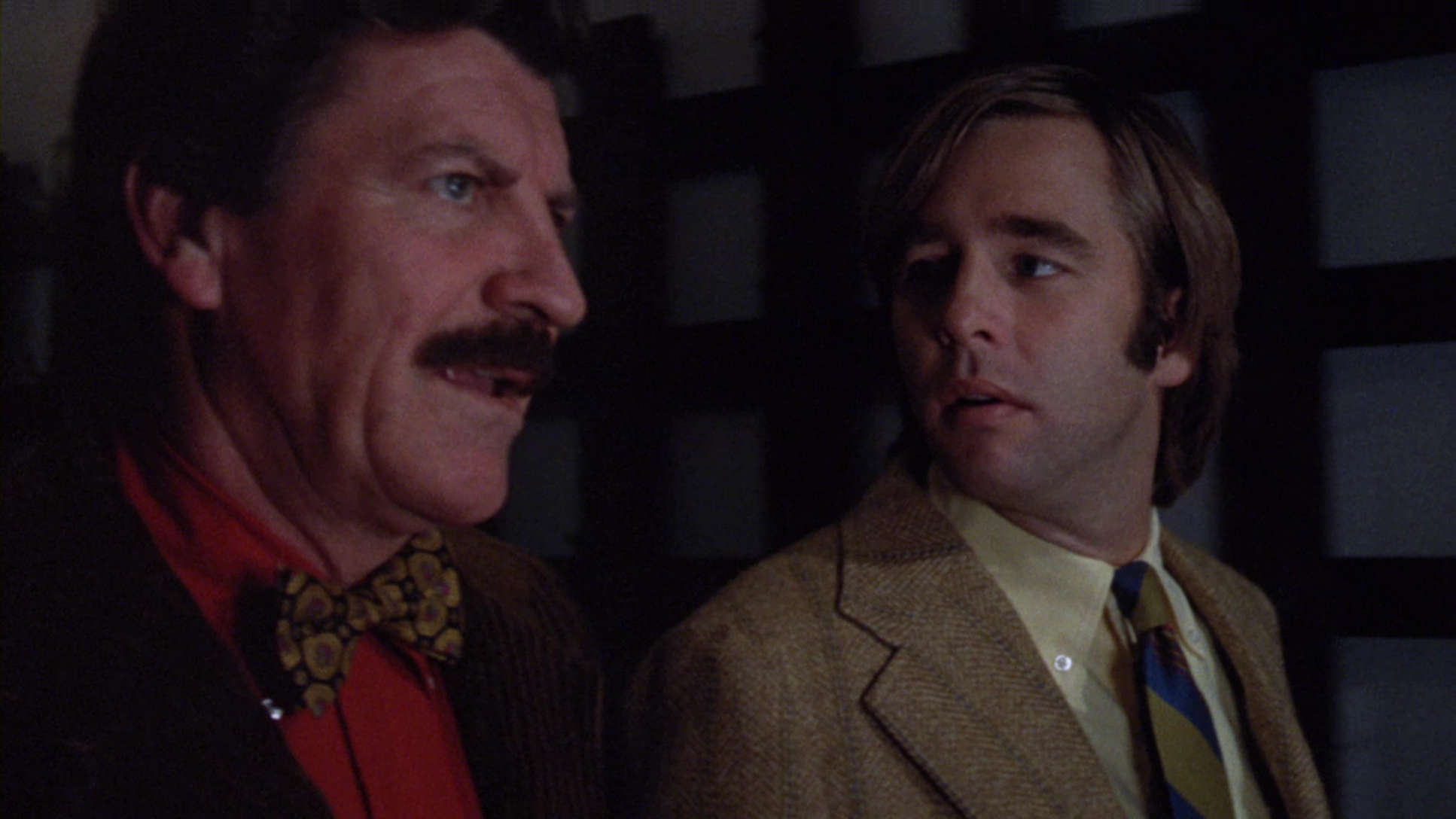 Two men standing side by side, with the man on the left sporting a mustache and wearing a red jacket with a bow tie, and the man on the right wearing a beige jacket and a striped.